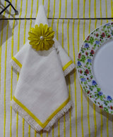 White cloth napkin with yellow embroidered border and fringed edges, held by a yellow flower-shaped napkin ring with a pearl center, on a yellow-striped tablecloth beside a floral-patterned plate.