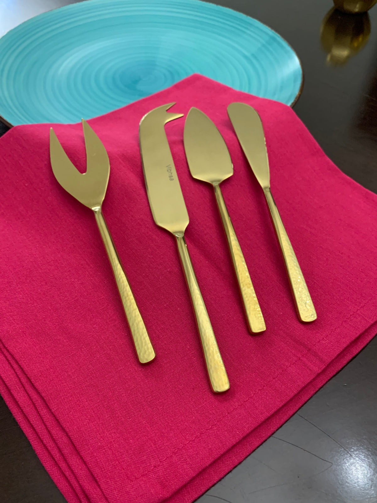 Set of four gold-toned stainless steel cheese knives with hammered handles, arranged on a red cloth near a blue ceramic plate on a dark surface.