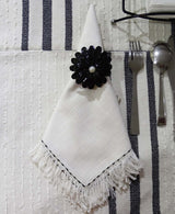 White cloth napkin with black embroidered edge and fringe ends folded into a triangle, held by a black flower-shaped napkin ring with a central pearl bead, placed on a white textured tablecloth with black stripes alongside silver,