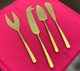 Set of four hammered stainless steel gold-tone cheese knives with distinct blade shapes, arranged on a bright pink cloth on a dark surface.