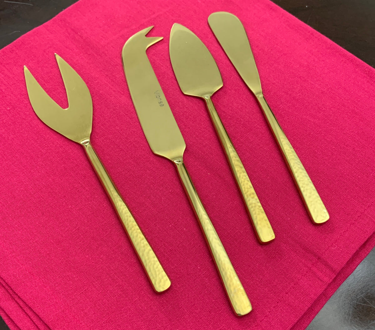 Set of four hammered stainless steel gold-tone cheese knives with distinct blade shapes, arranged on a bright pink cloth on a dark surface.