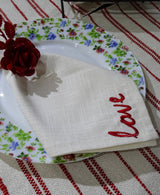 Cream-colored cloth napkin with red embroidered word 'love' placed on floral-rimmed plate, accompanied by a red decorative rose on striped beige and red tablecloth.