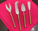 Set of four stainless steel cheese knives with gold twisted handles laid on a bright pink cloth, varying in blade shape for different cheese types.