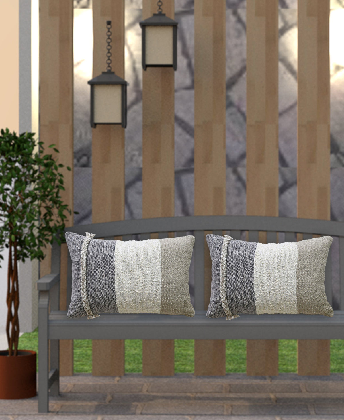 Gray wooden bench outdoors with two rectangular striped throw pillows in shades of cream, beige, and gray, flanked by a potted green plant and hanging lanterns.