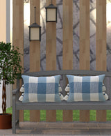 Gray outdoor bench with two textured blue and white plaid pillows featuring tassels on the sides, set against vertical wooden slats and flanked by lanterns and a potted plant.