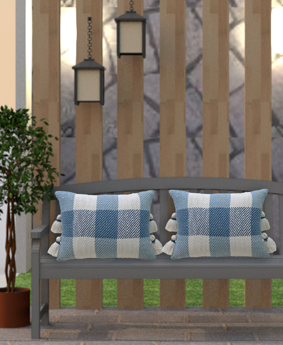Gray outdoor bench with two textured blue and white plaid pillows featuring tassels on the sides, set against vertical wooden slats and flanked by lanterns and a potted plant.