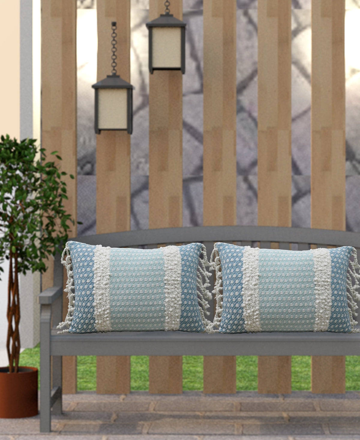 Two rectangular light blue pillows with white handwoven buttknot edging and fringe on a gray outdoor bench under hanging lanterns, against vertical wood slats.