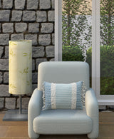 Light blue textured armchair with a rectangular decorative pillow featuring blue and white stripes and fringed edges, set beside a stone wall and tall lamp with green leaf pattern shade.