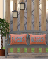 Two rectangular coral pillows with blue tassels and the word BLESSED embroidered in blue, placed side by side on a gray wooden bench outdoors.