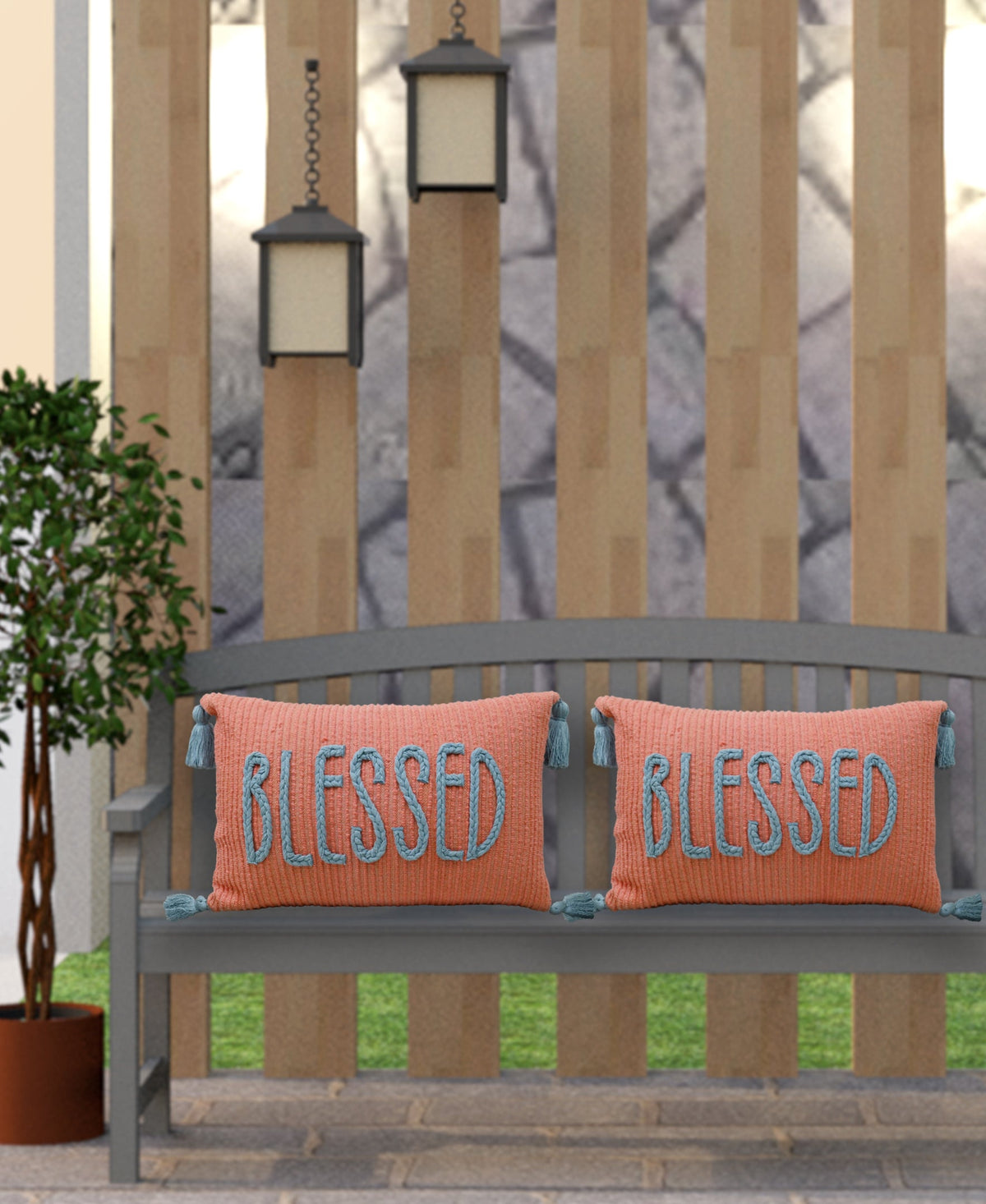 Two rectangular coral pillows with blue tassels and the word BLESSED embroidered in blue, placed side by side on a gray wooden bench outdoors.