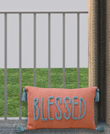 Rectangular coral-colored pillow with blue braided letters spelling BLESSED and blue tassels on each corner, shown against a gray railing and green grass background.