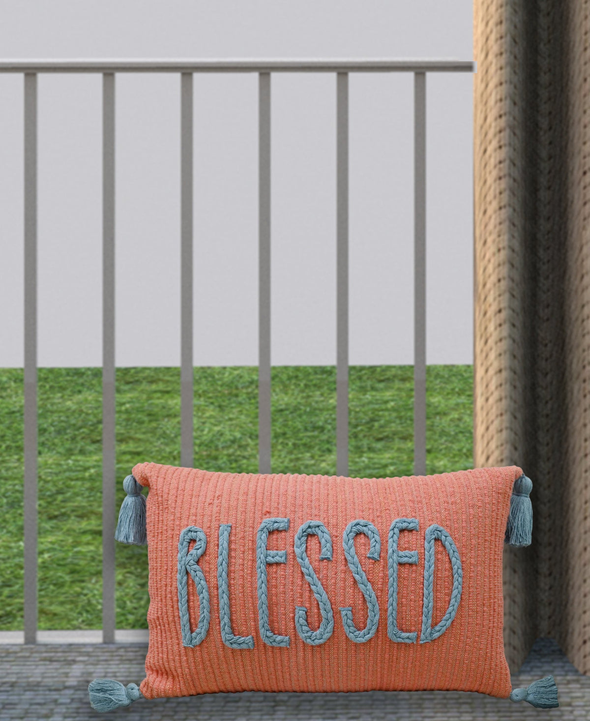 Rectangular coral-colored pillow with blue braided letters spelling BLESSED and blue tassels on each corner, shown against a gray railing and green grass background.