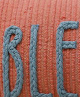 Close-up of braided blue yarn forming letters on a textured coral fabric background with horizontal weave patterns.