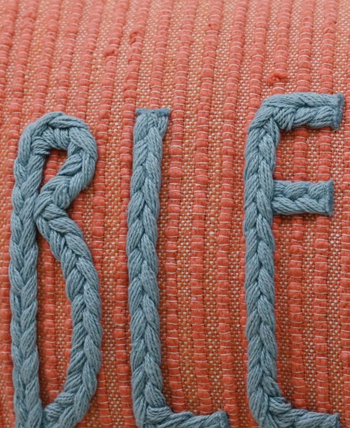 Close-up of braided blue yarn forming letters on a textured coral fabric background with horizontal weave patterns.