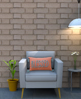 Light gray armchair with wooden legs holding a rectangular orange pillow embroidered with 'BLESSED' in blue, set against a brown brick wall, flanked by a green potted plant and a side table with white flowers under a hanging white
