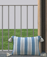 Rectangular pillow with blue and white vertical stripes and white tassels on each corner, displayed on a tiled floor against a railing with grass beyond.