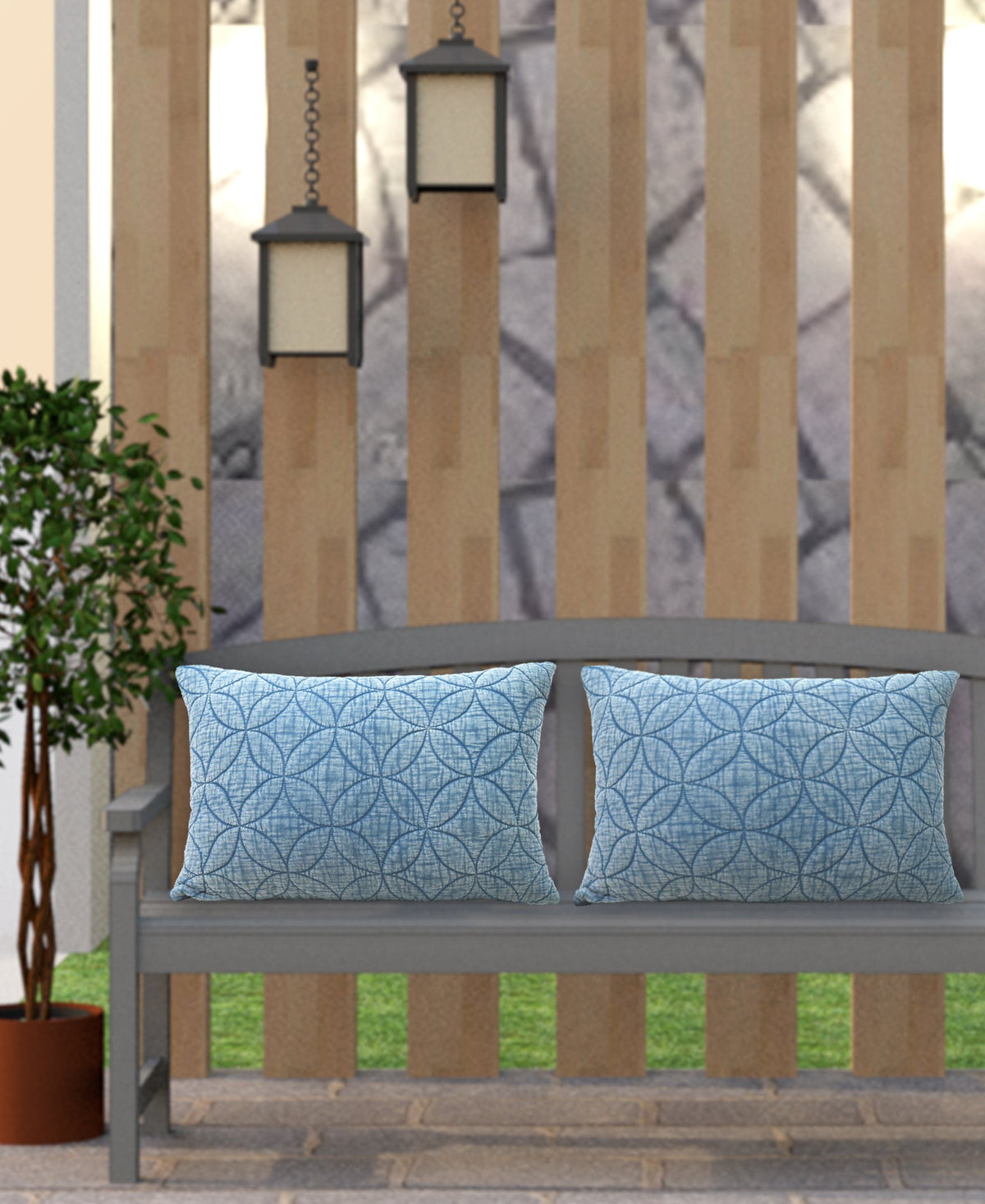 Two rectangular blue pillows with textured quilted circular pattern resting on a gray wooden bench against a vertical wood slat fence with two hanging lanterns and a potted plant nearby.