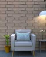 Light gray armchair with wooden legs holding a rectangular, textured quilted pillow featuring a blue geometric pattern, set against a beige brick wall with a potted plant and side table nearby.