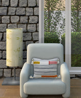 Light blue textured armchair with a rectangular decorative pillow featuring striped embroidery in gray with red, orange, and yellow accents and tassels on corners, placed beside a lamp with a leafy patterned shade against a stone-