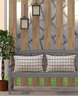 Two rectangular decorative throw pillows with orange knotted edging and white grid pattern on a gray outdoor bench against a wooden slat backdrop with two hanging lanterns.