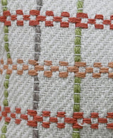 Close-up of woven fabric with a grid pattern of vertical green and gray lines intersecting horizontal orange and rust-colored knotted stripes on a beige background.