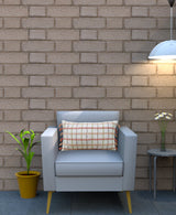 Light gray armchair with a rectangular throw pillow featuring orange handwoven knotted edging and circular patterns, set against a beige brick wall with a hanging lamp and nearby plant.