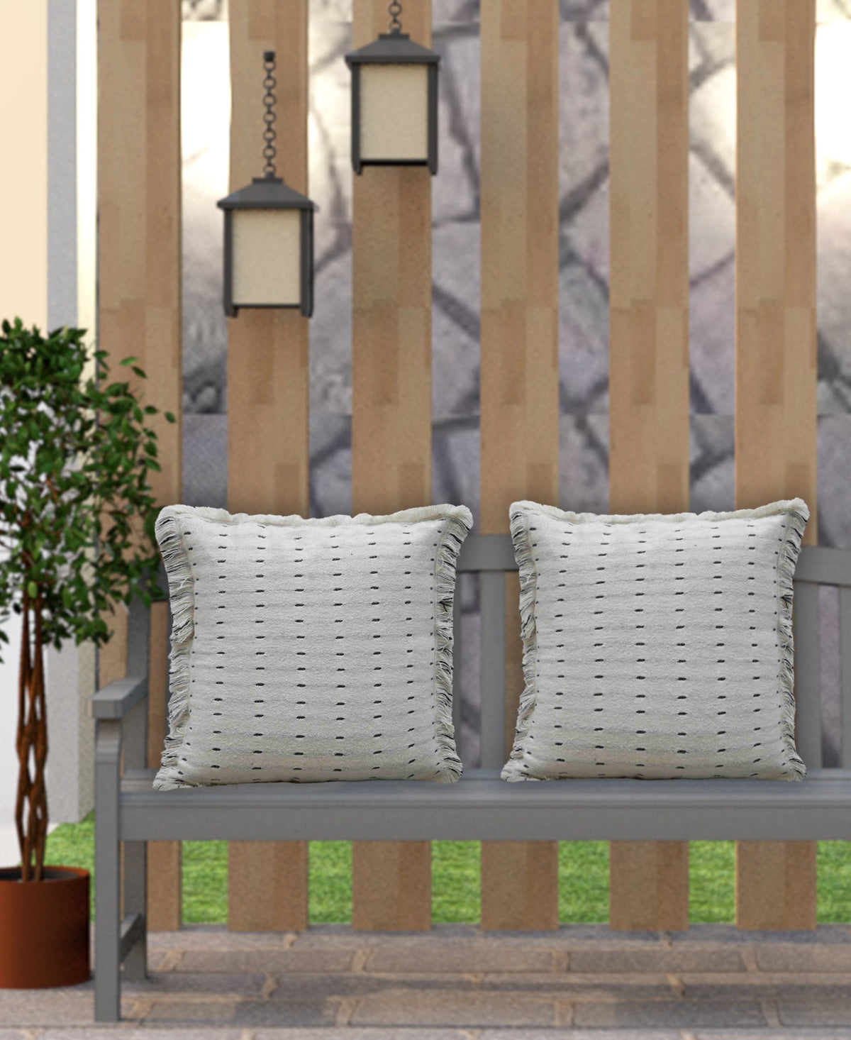 Two light gray decorative pillows with subtle black dotted pattern on a gray wooden bench against a vertical beige slatted fence with two hanging lanterns nearby.