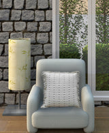 Soft textured light gray armchair with a textured white decorative pillow on it, positioned near a tall lamp with a beige shade on a stone wall background.