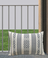 Rectangular beige throw pillow with black woven geometric diamond patterns and vertical stripes, resting on a gray floor with green grass and railing background.