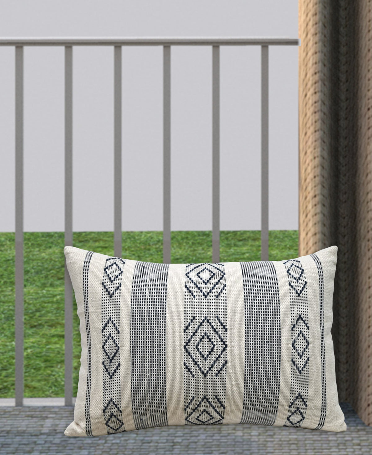 Rectangular beige throw pillow with black woven geometric diamond patterns and vertical stripes, resting on a gray floor with green grass and railing background.