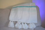 Hand-loomed white woven throw with turquoise geometric patterned stripes and large white tassels, draped over a textured beige couch armrest.