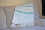 White woven throw with turquoise horizontal stripes and diamond patterns, accented with white tassels, draped over a light beige textured sofa armrest.