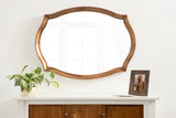 Scalloped wooden-framed wall mirror with smooth curves, hung above a white cabinet topped with a green potted plant and a framed photo.