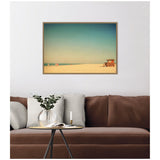 Framed wall art above a brown sofa, depicting a serene beach scene with a sandy shore, clear blue sky, and a small lifeguard hut on the right side.