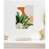 Framed abstract botanical print featuring green tropical leaves with geometric shapes in terracotta, beige, and light pink, hanging on a white wall above a minimal desk.