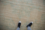 Worn vintage Oushak carpet in muted beige with faded blue-green detailing, viewed from above with a person wearing black and white sneakers standing on it.