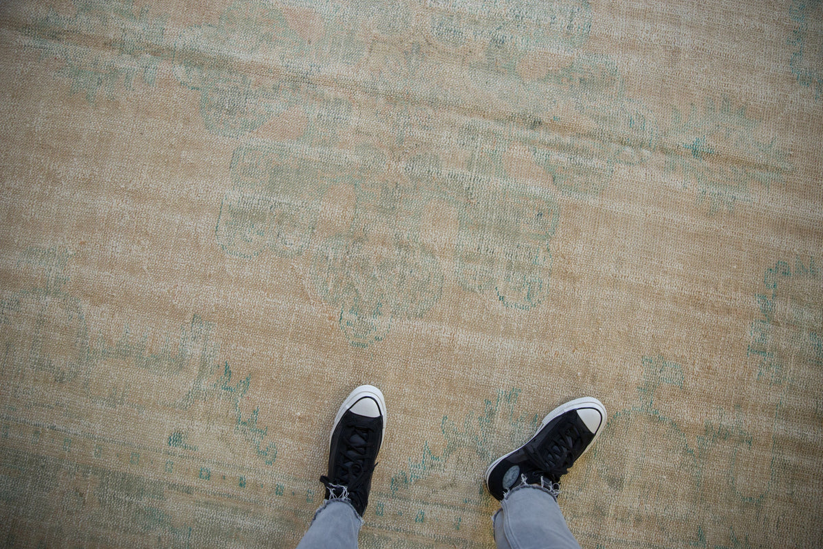 Worn vintage Oushak carpet in muted beige with faded blue-green detailing, viewed from above with a person wearing black and white sneakers standing on it.