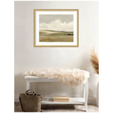 Neutral-toned framed landscape painting of rolling hills and cloudy sky above a white bench draped with a fluffy beige throw, next to a brown bag and pampas grass.