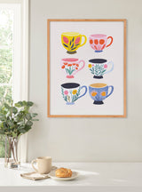 Framed print of six colorful illustrated floral coffee cups with pink, yellow, blue, and orange hues displayed on a light wall above a white table with a beige mug, croissant, and green plant.