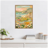 Framed canvas painting of a coastal landscape with colorful wildflowers, rolling hills, and calm sea under a soft pastel sky, displayed on a white wall.