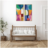 Two vertical abstract canvas panels with colorful geometric shapes in green, blue, orange, pink, and white, framed in light wood, hanging above a wooden bench with white cushions.