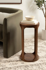 Tall, round Dahl wood drink table with a sculpted base and smooth finish, holding stacked books and a small bowl, placed beside a green velvet armchair on a patterned rug.