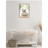 Framed canvas showing a baby elephant sitting in a vintage clawfoot bathtub with green palm leaves framing the scene, hung on a plain light wall above a white bench with a fur throw and a beige bag below.