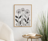 Black and white linocut botanical print with stylized flowers and leaves in a light wood frame hanging above a round brass side table and white bowl, next to a cream chair and tall grass plant.