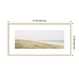 Framed wall art print depicting a peaceful coastal landscape with grassy dunes in soft green and a pale blue ocean under a light sky, bordered by a light natural wood frame with white matting.