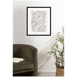 Framed artwork featuring an abstract continuous black line drawing on a light beige background, hanging above a dark olive curved armchair with an open book on the seat, next to a potted plant with green leaves and small dark red-