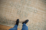 Person wearing black boots and blue jeans standing on a beige vintage distressed Oushak carpet with subtle faded floral and medallion patterns.