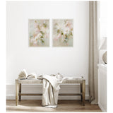 Two framed canvas prints featuring delicate pastel floral designs in soft pinks and greens, hung side by side on a white wall above a wooden bench with draped fabric.