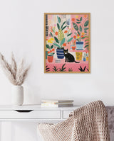Framed colorful print of various potted houseplants with a black cat sitting among them, displayed above a white desk with books and a vase of pampas grass.
