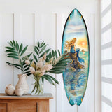 Surfboard wall art depicting a mermaid with long flowing hair sitting on rocks at sunset, surrounded by calm ocean waters in soft blue and orange hues.